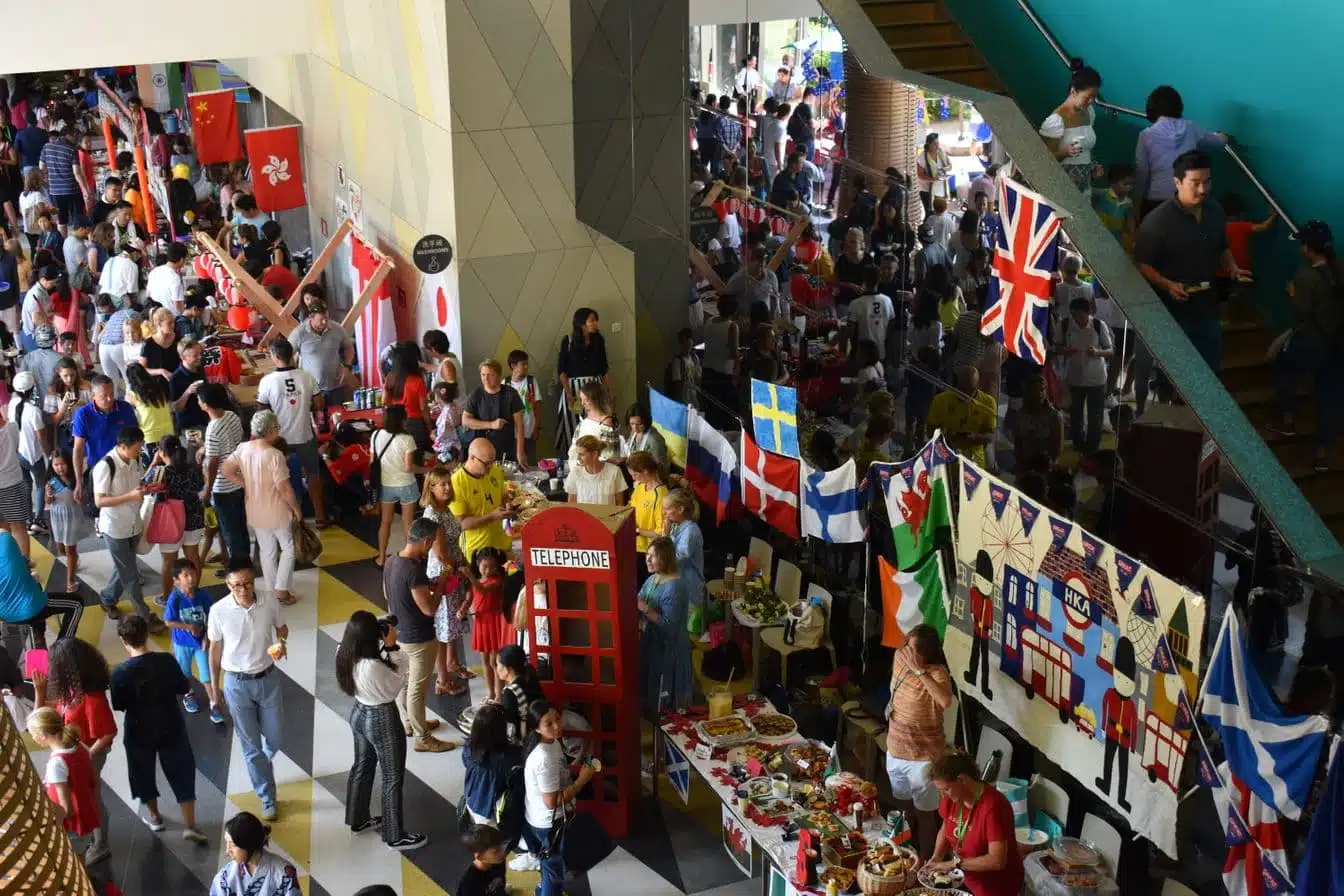 Families and students celebrating cultural diversity at a Hong Kong Academy community event featuring international flags and food stalls in our Sai Kung campus atrium.